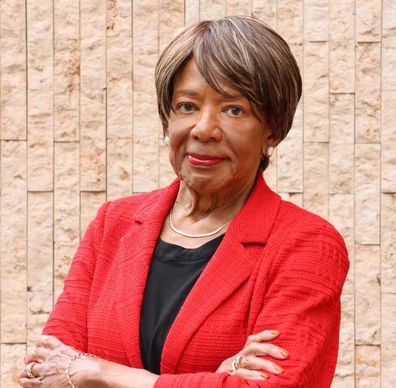 Deborah Richardson standing in front of a light brown brick wall, wearing a red jacket, arms crossed